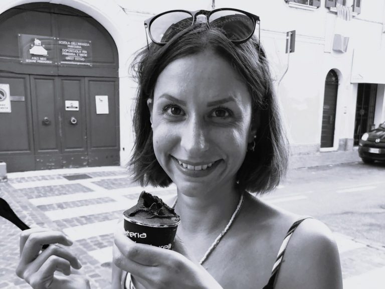 Alina Jessl mit Kurzhaarfrisur, die ein Dessert lächeln hält, auf einer Straße.