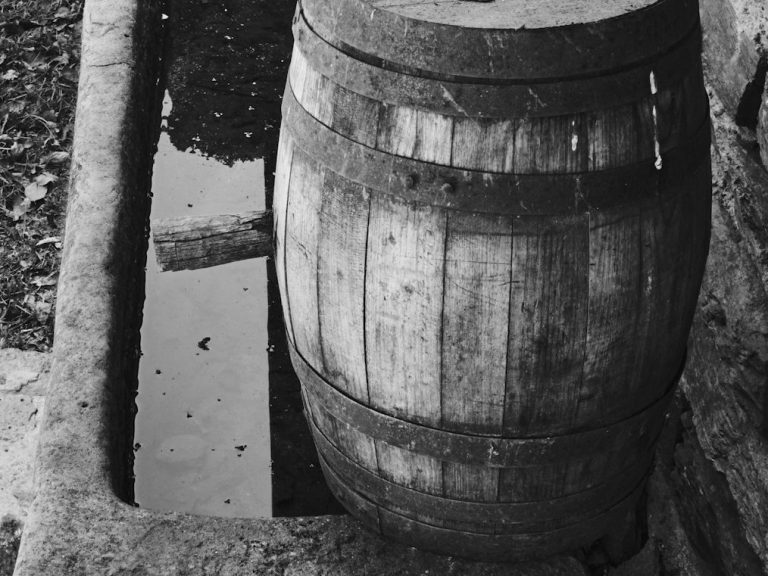 Ein Holzfass steht neben einer schmalen, flachen Wasserstelle.