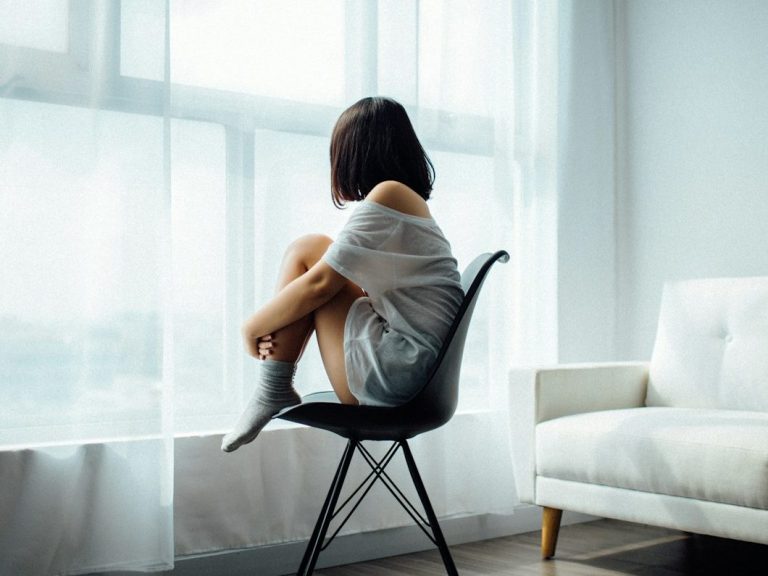 Eine Person sitzt auf einem Stuhl und blickt aus einem Fenster in einen hellen Raum.