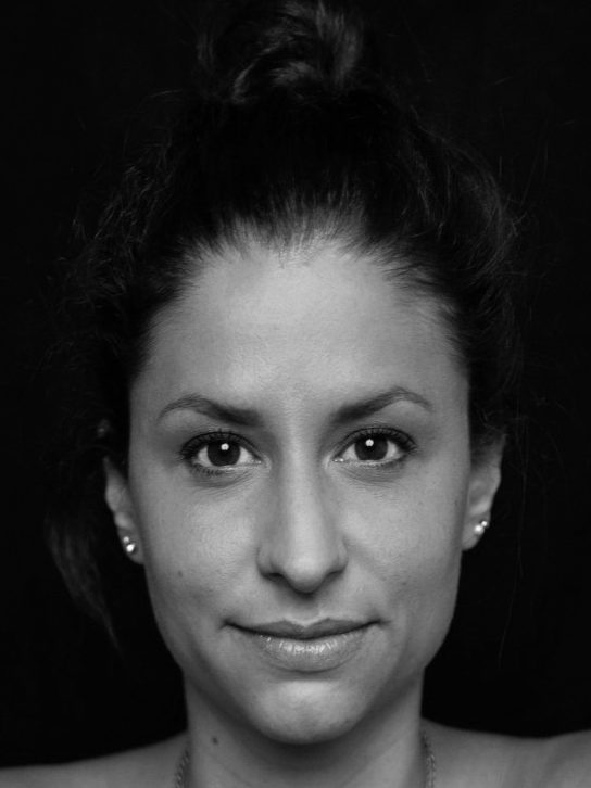 Porträt Alina Jessl mit dunklem Haar als Dutt, lächelnd und  vor schwarzem Hintergrund. Die Abbildung zeigt nur den Kopf, ist schwarz weiß