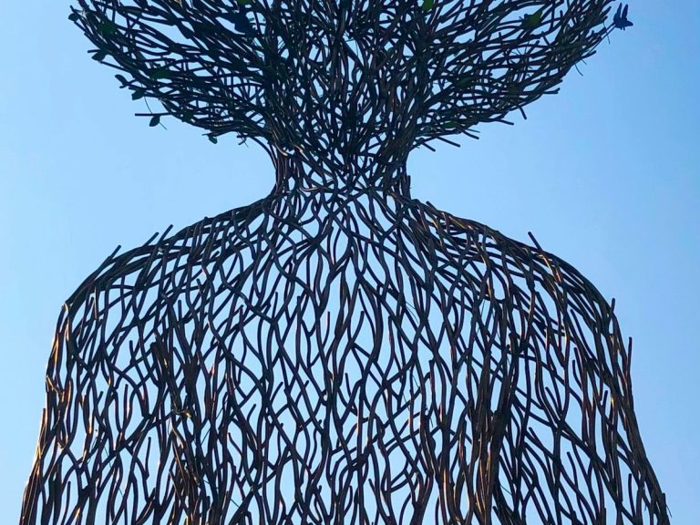 Sculpturale Darstellung einer menschlichen Figur aus verflochtenen Ästen vor blauem Himmel.