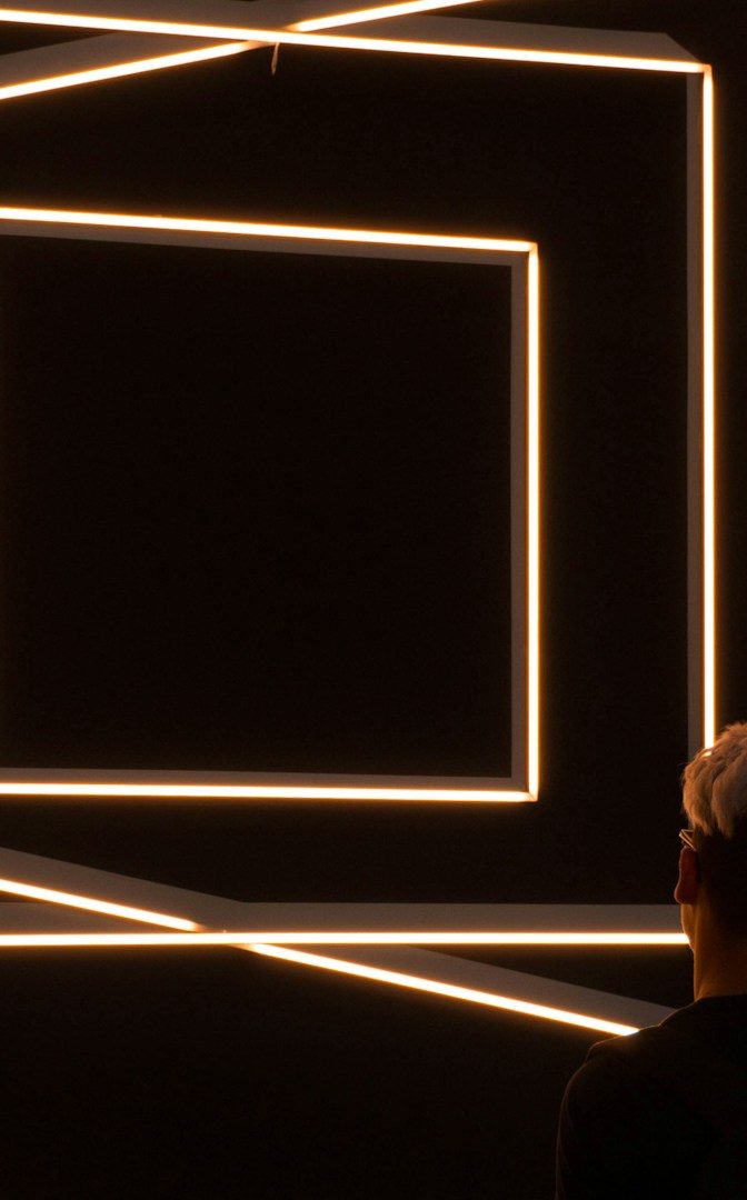 Person steht vor einer Wand mit leuchtenden, geometrischen Linien in einem dunklen Raum.
